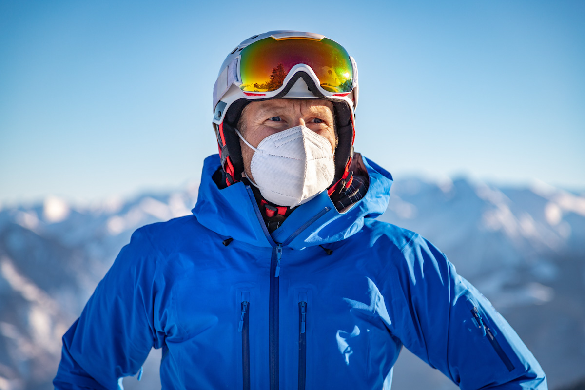 Ein Mann in einer blauen Jacke in winterlich-verschneiten Bergen trägt eine FFP2-Maske.