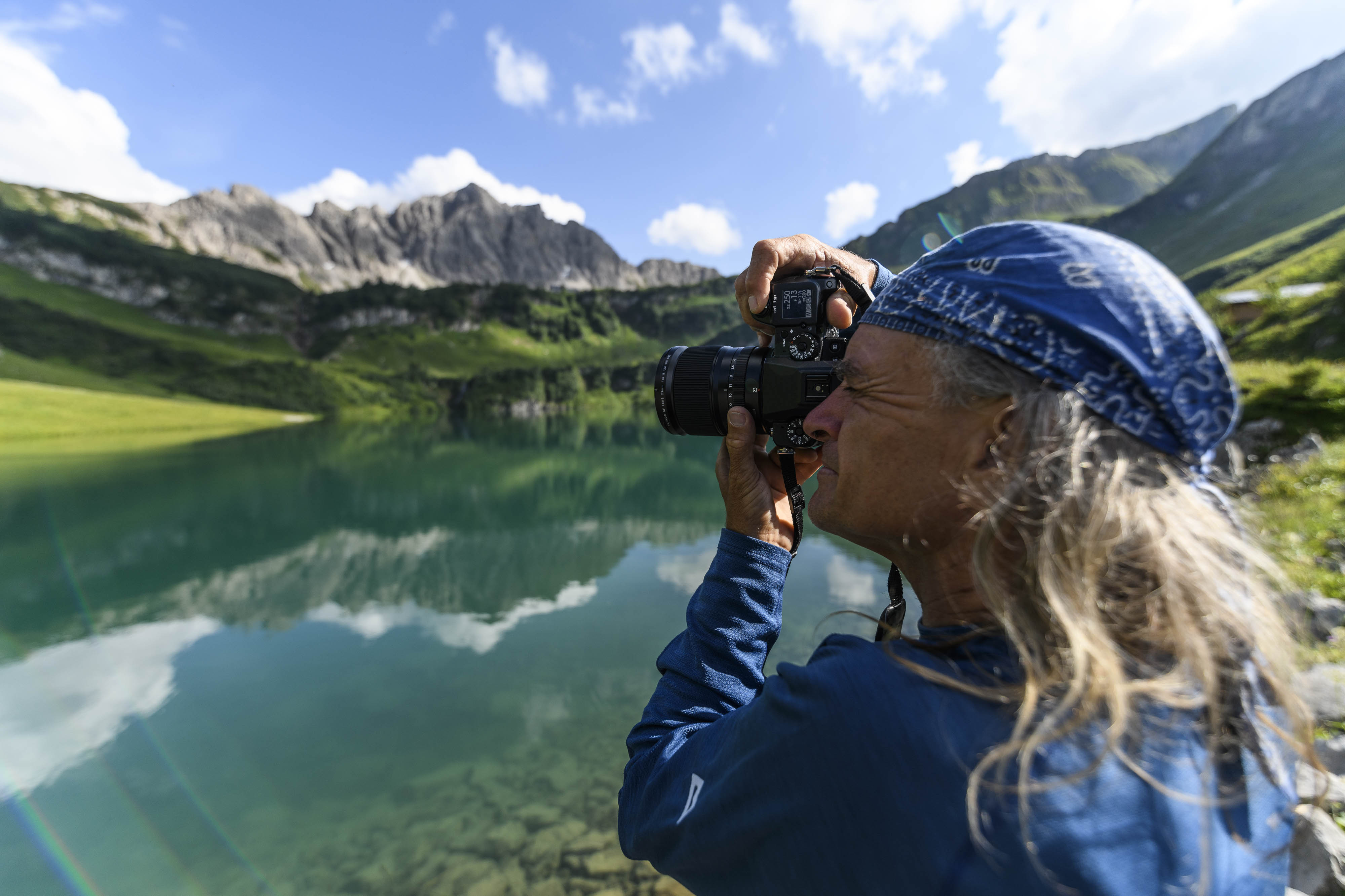 Der Fotograf Heinz Zak steht an einem Bergsee und schaut durch seine Spiegelreflexkamera.